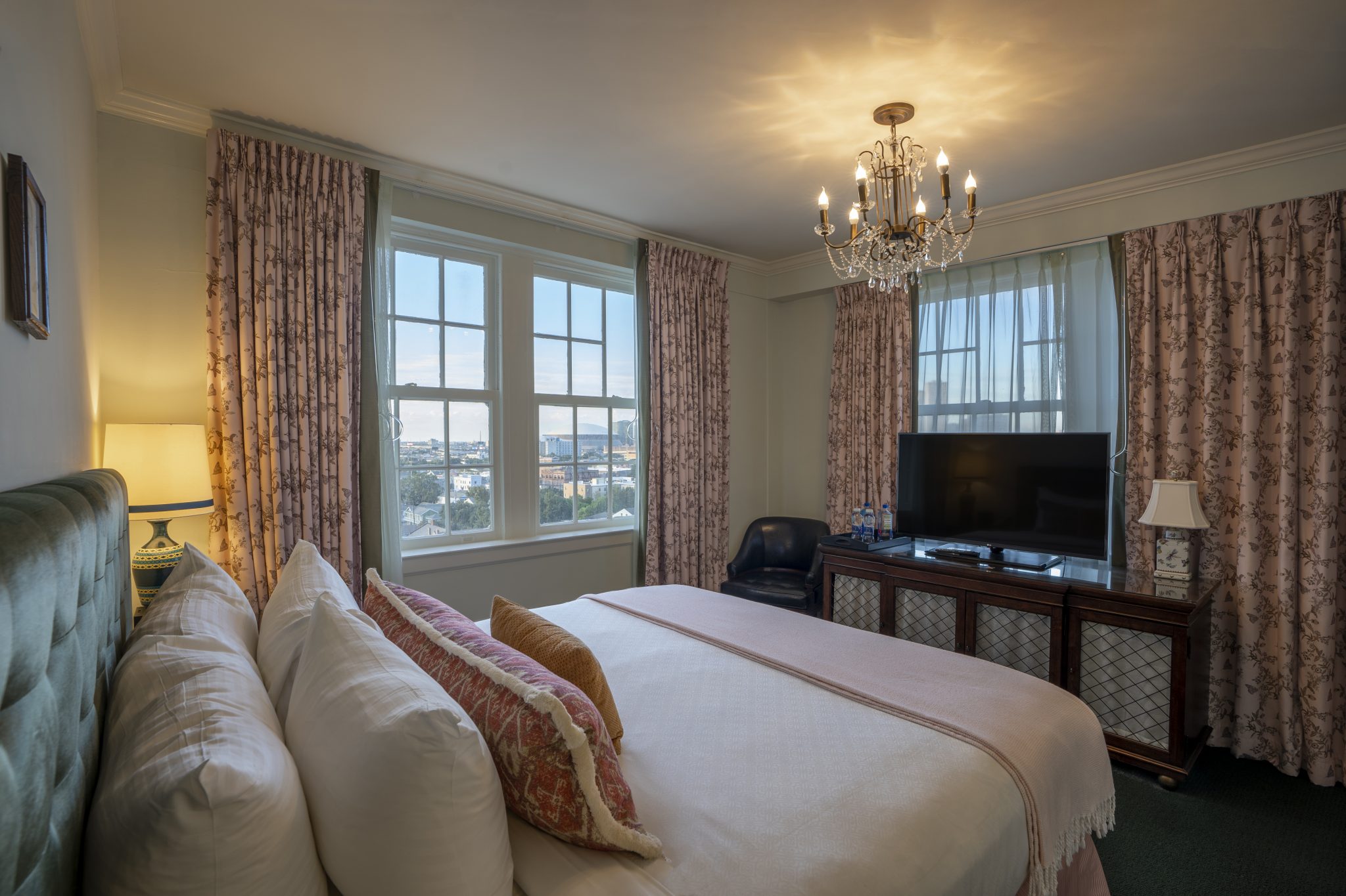A wide-angle view of a hotel room with a bed, TV, chandelier, and window.